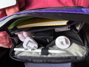 A purple tech pouch, filled with plugs, an Apple Pencil, a pink UFO looking thing, a small notebook and some plastic camping cutlery.